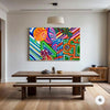 A large, vibrant abstract canvas print with multicolored patterns hangs on a white wall above a wooden dining table and bench in a modern, sunlit room.