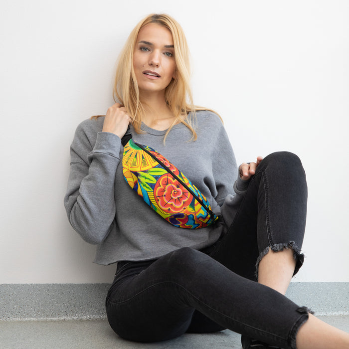 A blonde woman sitting on a gray floor against a white wall, wearing a gray sweatshirt and black jeans, with a colorful floral embroidered fanny pack slung across her chest.