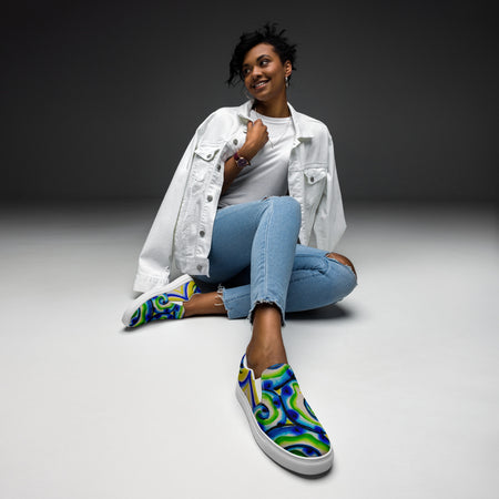 woman sitting wearing  slip on canvas shoes with all over multicolored print in swirling blues and greens