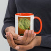 A person holding a white ceramic mug with a bright orange interior and handle. The front of the mug features a printed illustration of a green artichoke over orange wavy stripes.