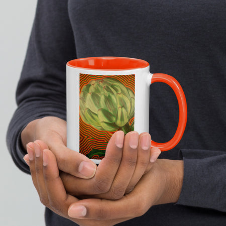 A person holding a white ceramic mug with a bright orange interior and handle. The front of the mug features a printed illustration of a green artichoke over orange wavy stripes.