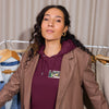 A woman with long curly dark hair is wearing a brown blazer over a burgundy hoodie, standing in front of a clothing rack with other garments.