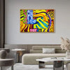 A colorful abstract canvas wall art print with bold yellow, blue, green, pink, and red patterns, displayed in a modern living room setting above a beige sofa.