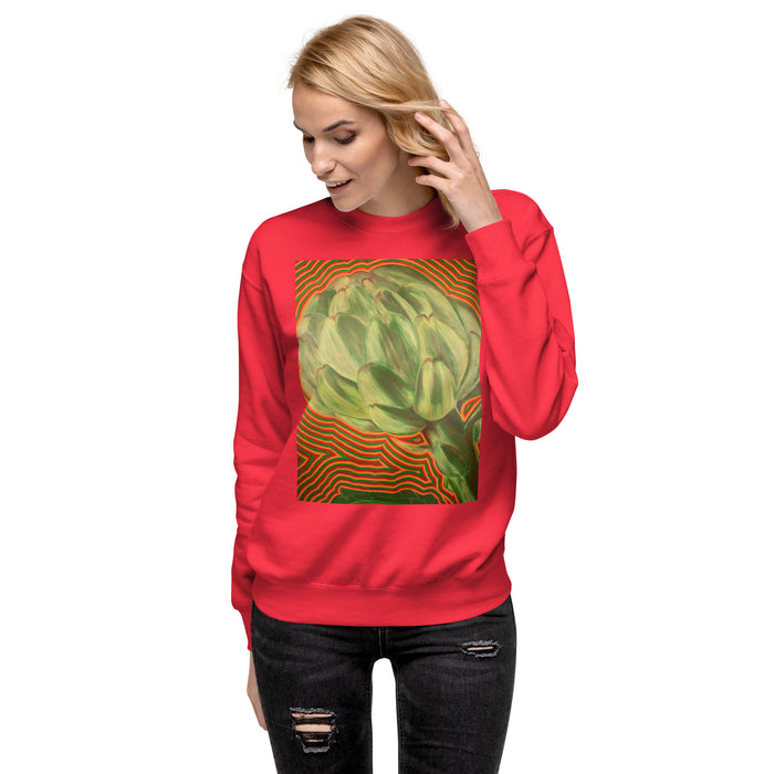 A woman modeling a bright red crewneck sweatshirt featuring a large graphic print of a green artichoke on a background of orange and green wavy lines. She is wearing dark distressed jeans against a solid white studio background.