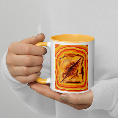 A person in a white long-sleeve shirt holds a white ceramic mug with a yellow interior and handle. The mug features a colorful graphic print of a toasted slice of bread with a diagonal cut, surrounded by concentric orange and yellow rings.