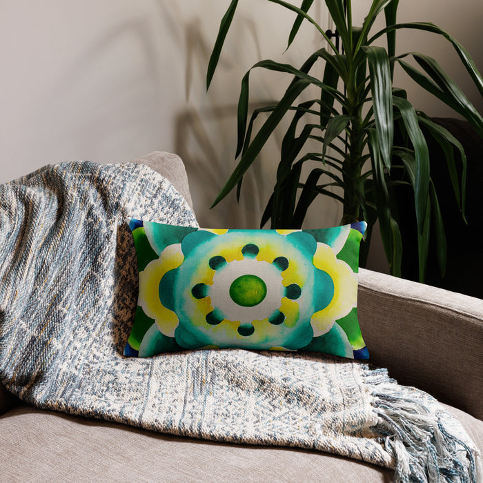 A rectangular lumbar throw pillow with a vibrant green, yellow, and blue mandala pattern, styled on a beige armchair draped with a textured blue and white knit throw blanket, with a green houseplant in the background.