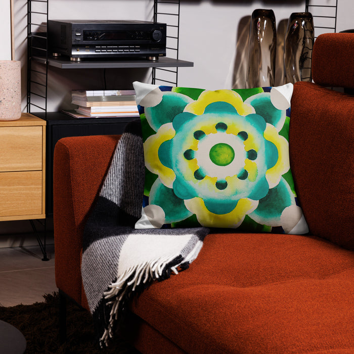 A square throw pillow with a vibrant green, yellow, and teal mandala pattern, styled on a rust-orange armchair next to a black-and-white plaid throw blanket in a modern living room setting.