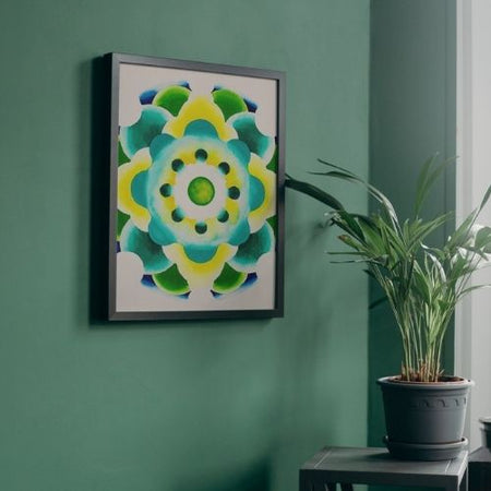 Framed wall art featuring a colorful abstract floral mandala design in shades of green, yellow, and blue, hung on a dark green wall next to a potted plant.