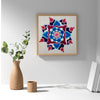 A square framed wall art print featuring a colorful mandala design in shades of blue, red, and pink on a white background, styled on a white shelf next to a vase with greenery and books.
