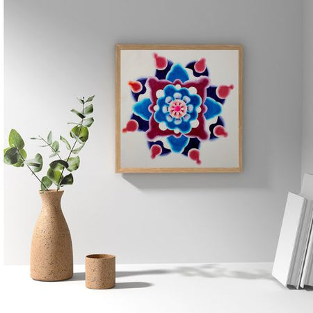 A square framed wall art print featuring a colorful mandala design in shades of blue, red, and pink on a white background, styled on a white shelf next to a vase with greenery and books.