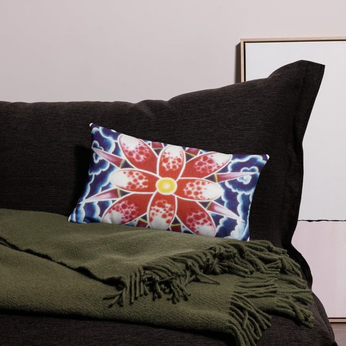 A rectangular lumbar throw pillow with a vibrant red, white, and blue floral mandala pattern, styled on a dark brown sofa next to an olive green fringed throw blanket.