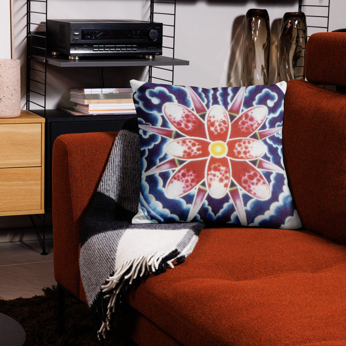 A square throw pillow with a colorful mandala-style floral pattern in red, white, blue, and yellow, resting on an orange-brown sofa next to a black-and-white plaid throw blanket.
