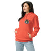 A woman modeling a coral orange pullover hoodie with a circular bardo kid graphic on the left chest, paired with light blue jeans against a plain white background.