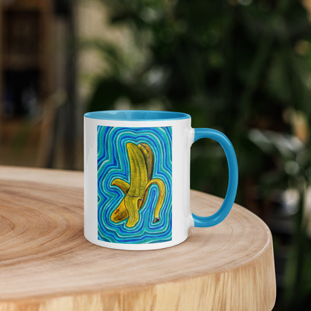 A white ceramic mug with a bright blue handle and interior, featuring a graphic print of a yellow banana peel on a swirling blue and green patterned background, sitting on a wooden table with a blurred green garden background.