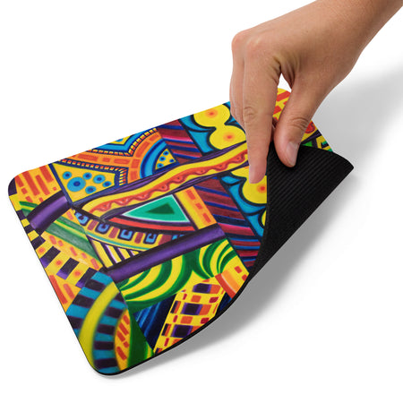 A hand is lifting the corner of a rectangular mouse pad to show its black textured underside. The top surface features a vibrant, multicolored abstract geometric pattern with yellow, green, blue, purple, and red shapes.